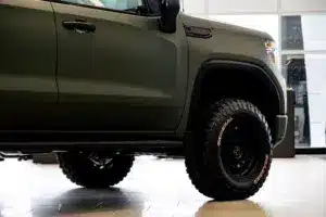 Side view of a rugged truck with an olive green exterior and large off-road tires, displayed in a modern showroom environment with bright lighting and sleek flooring.