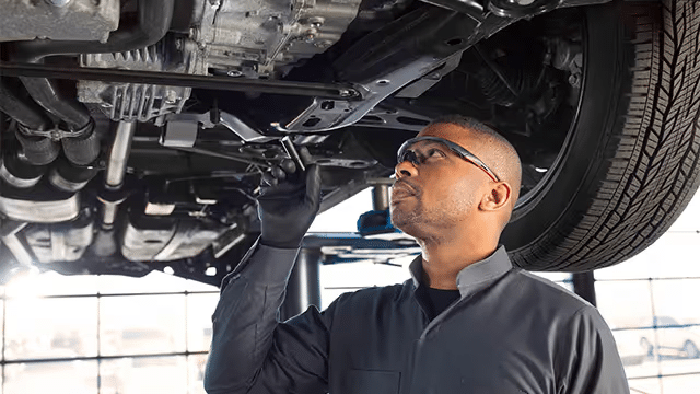 Mechanic inspecting the underside of a vehicle while wearing protective gloves and glasses in an automotive workshop.