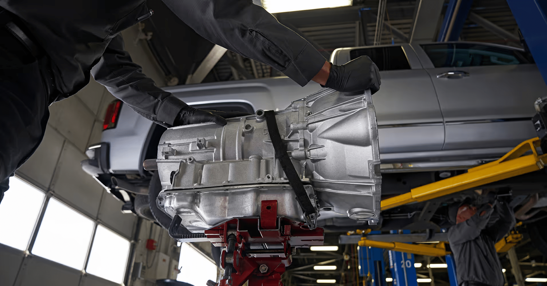 Mechanic holding a silver transmission ready for installation under a vehicle in an auto repair shop, with another technician working on the car in the background.