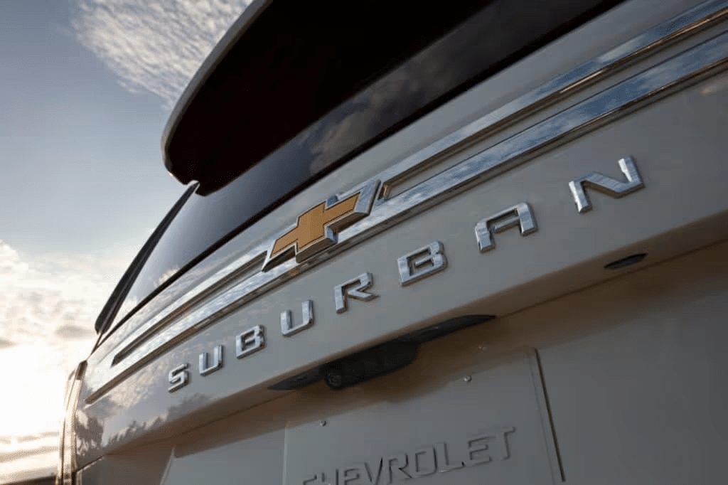 Close-up view of the rear of a Chevrolet Suburban, highlighting the chrome logo and lettering against a scenic sky backdrop, showcasing the vehicle's design and brand.