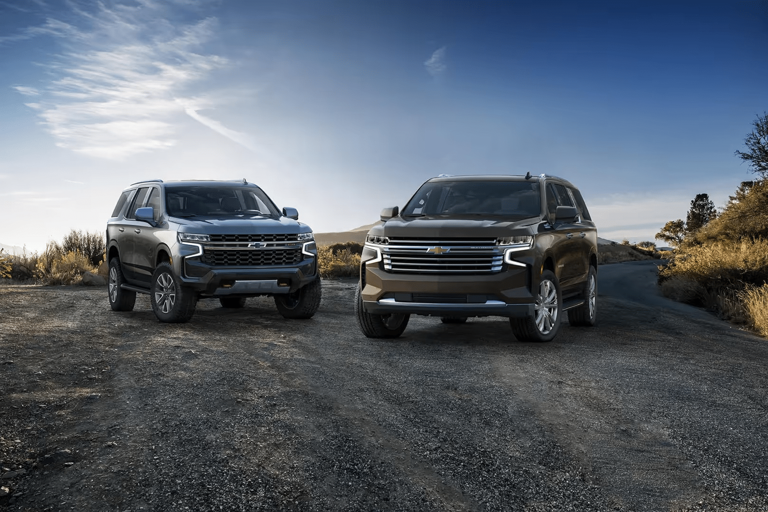Two Chevrolet SUVs parked on a gravel road, showcasing a blue and a brown model against a scenic backdrop of sky and hills. The image highlights their modern design and rugged features, ideal for outdoor adventures.