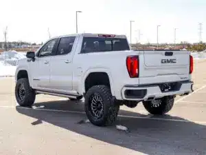 White GMC Denali truck parked in an empty lot, displaying its rugged design and large Black wheels. The image captures the vehicle's rear view, showcasing its distinctive tail lights and brand logo, set against a clear sky and snow-covered ground.