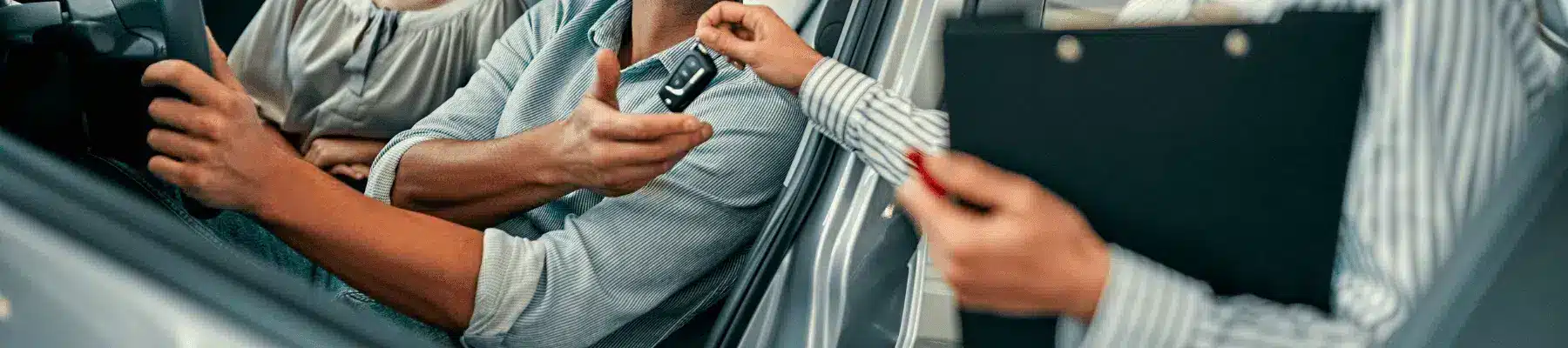 A man in a car receives a set of keys from a woman while another person observes, illustrating a car rental or leasing transaction in a professional setting.