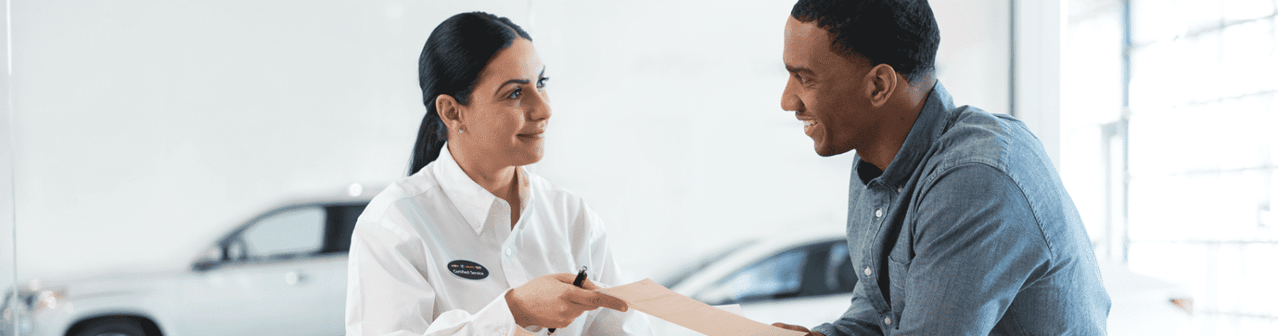 Customer service representative assisting a client in a dealership, discussing paperwork with a friendly demeanor; a vehicle is visible in the background.