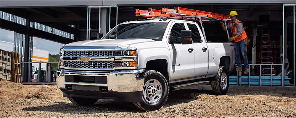 White Chevrolet Silverado pickup truck with ladder rack parked at a construction site, featuring a construction worker in safety gear preparing to load materials.