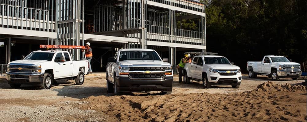 Four Chevrolet pickup trucks are parked at a construction site, with workers nearby preparing for a project. The vehicles are positioned in a sandy area in front of a partially constructed building, showcasing their utility in a work environment.