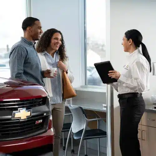Couple receiving customer service at a Chevrolet dealership, with a salesperson reviewing details on a tablet next to a red Chevrolet vehicle. Bright, modern showroom ambiance.