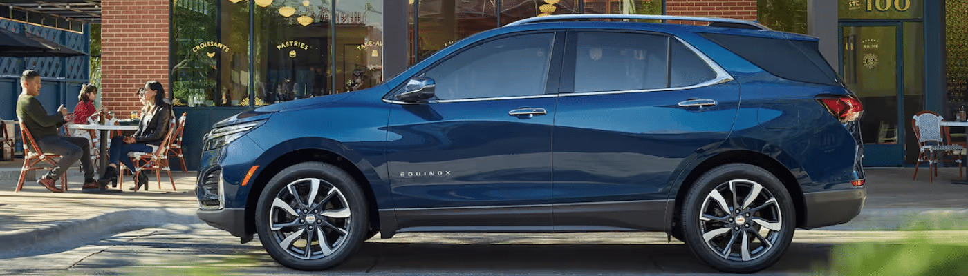 Side view of a blue Chevrolet Equinox parked near a café, showcasing its modern design and sleek lines, with people enjoying outdoor seating in the background.