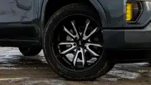 Close-up of a modern vehicle's black alloy wheel featuring a sleek design with silver accents, mounted on a tire with visible tread. The setting shows wet ground, indicating potential off-road use or recent weather conditions.