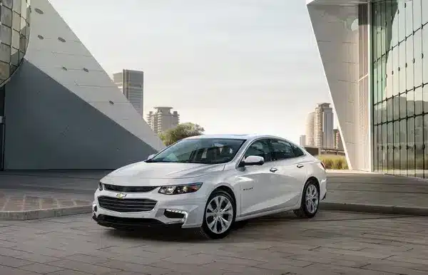 White Chevrolet Malibu parked in an urban setting with modern architecture in the background, showcasing its sleek design and stylish features.