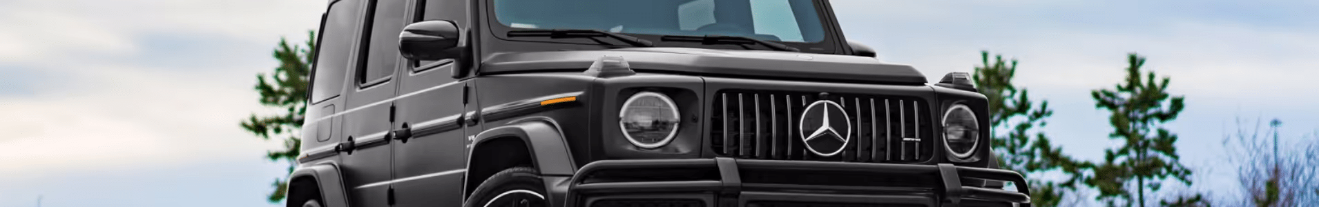 Close-up view of a black Mercedes-Benz G-Class SUV against a cloudy sky and trees, showcasing its iconic design and luxury features.