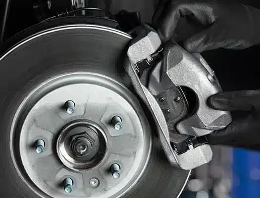 Close-up of hands wearing black gloves holding a metallic brake caliper next to a silver brake disc with lug nuts visible. The image shows part of a vehicles wheel assembly.