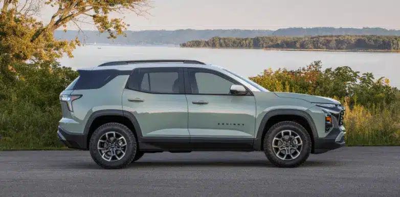 Side view of a green 2023 Chevrolet Equinox SUV parked by a scenic lake, surrounded by trees and hills, showcasing its modern design and off-road capabilities.
