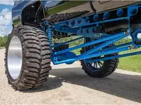 Detailed view of a modified truck’s suspension system, showcasing blue components and oversized tires, set against a nature backdrop. Ideal for off-road performance and customization enthusiasts.