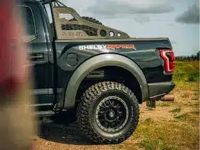 Close-up view of a black Shelby F-150 truck, showcasing its rugged tires, custom wheel design, and distinctive rear branding in an outdoor setting.