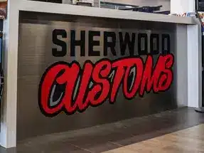 Signage displaying the logo of Sherwood Customs, featuring bold red lettering on a metallic background, located in a modern retail space.