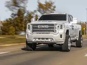 Powerful GMC truck driving on a scenic road, showcasing its sleek design and off-road capabilities against a backdrop of autumn foliage.