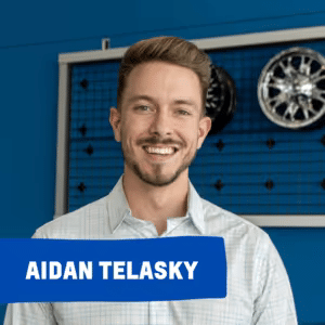 Smiling man in a light blue shirt standing in front of a display featuring automotive wheels, showcasing Aidan Telasky.