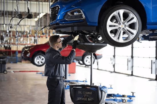 Mechanic performing an oil change on a blue car lifted above a service bay in an automotive workshop, with a red vehicle visible in the background.