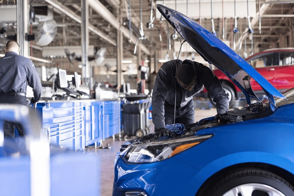 Mechanic performing an oil change on a blue car lifted above a service bay in an automotive workshop, with a red vehicle visible in the background.