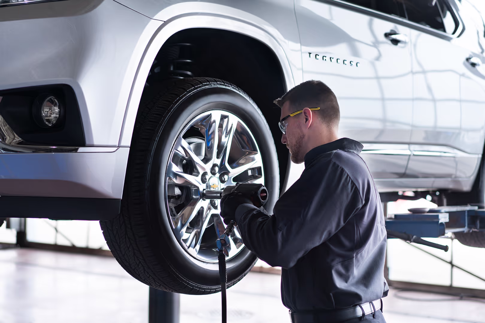 Mechanic performing an oil change on a blue car lifted above a service bay in an automotive workshop, with a red vehicle visible in the background.