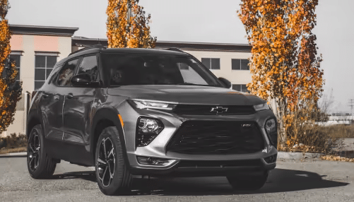 Silver 2023 Chevrolet Trailblazer SUV parked in front of a modern building with autumn trees in the background, showcasing its sleek design and sporty features.