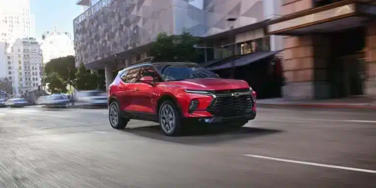 Red Chevrolet Blazer SUV driving through a city street with modern architecture and blurred traffic in the background, showcasing its sleek design and performance.