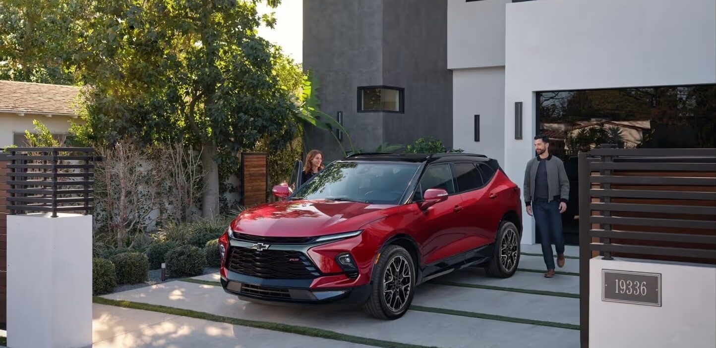 A couple stands near a red Chevrolet Blazer parked in front of a modern home, surrounded by greenery. The scene showcases the sleek design of the vehicle and the contemporary architecture of the house, emphasizing a stylish outdoor lifestyle.