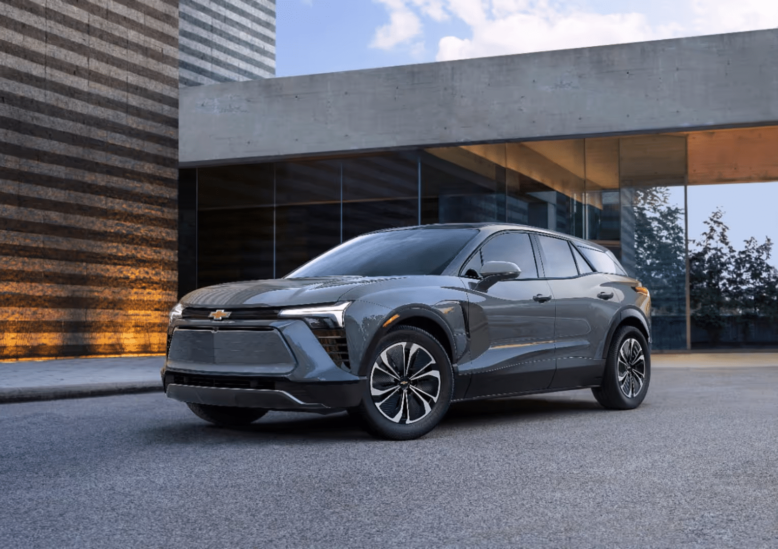 A sleek grey Chevrolet SUV parked in front of a modern architectural building with large glass windows and textured stone walls, showcasing contemporary design and automotive innovation.