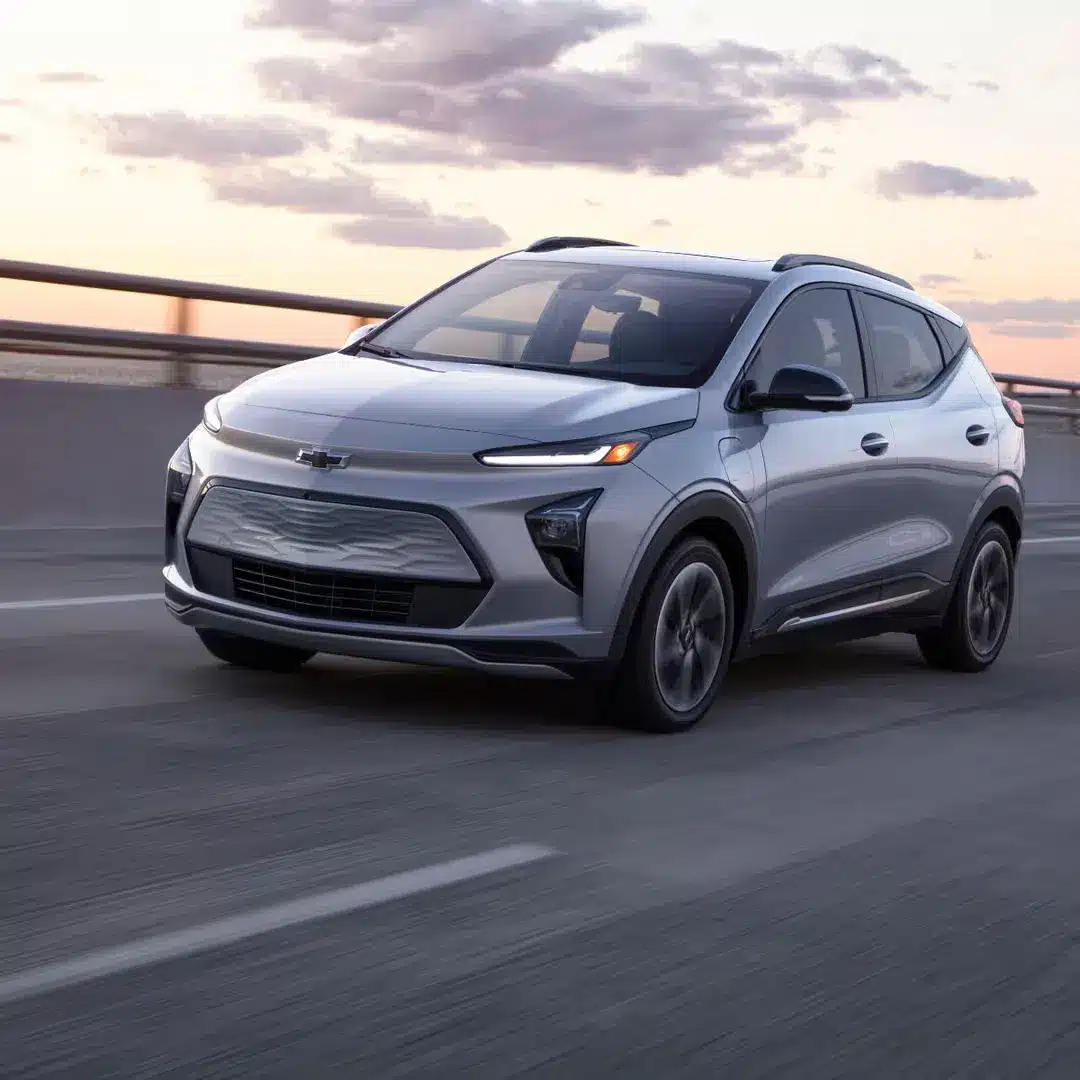 Silver Chevrolet electric SUV driving on a scenic highway during sunset, showcasing modern design and environmentally friendly features.