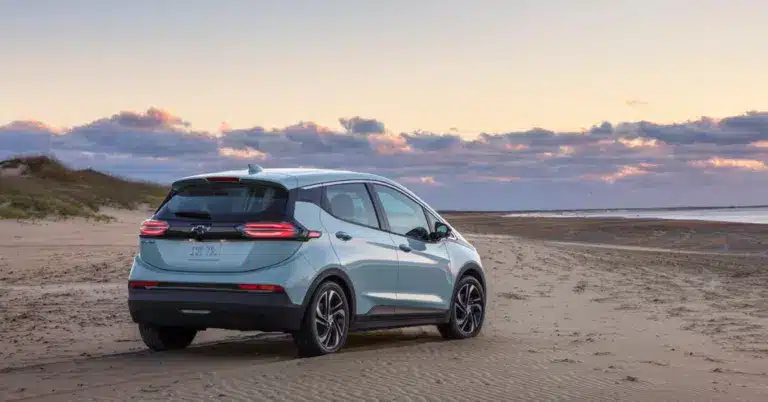 Electric Chevrolet Bolt EV parked on a sandy beach at sunset, showcasing its sleek design against a backdrop of clouds and coastline.