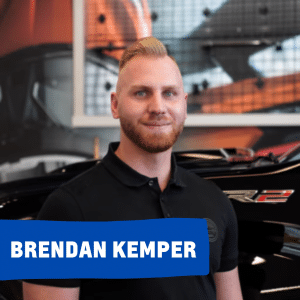Image of a man named Brendan Kemper standing in front of a black truck with a vibrant background. He is wearing a black polo shirt and smiling, showcasing a professional atmosphere at a truck dealership or automotive service center.