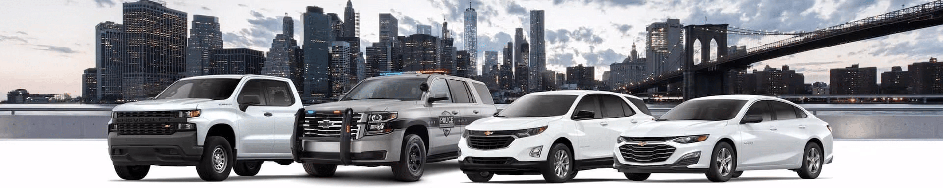 Chevrolet vehicles lineup featuring a pickup truck, police SUV, compact SUV, and sedan parked in front of a city skyline with modern skyscrapers.