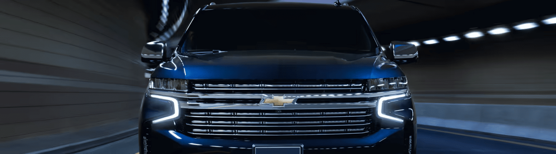 Front view of a blue Chevrolet truck driving through a dimly lit tunnel, showcasing its modern design and distinctive grille. The image emphasizes the vehicle's sleek lines and advanced lighting features, perfect for advertisements or automotive reviews.