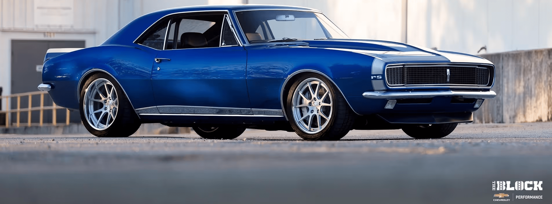 Classic blue Chevrolet Camaro RS parked with a sleek profile, featuring modern alloy wheels and a polished finish, set against an industrial backdrop. Ideal for automotive enthusiasts and classic car fans.