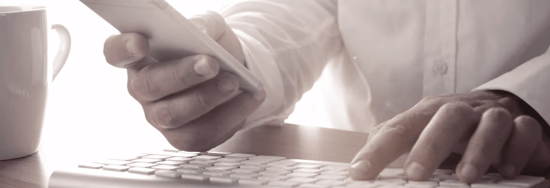 Close-up of a person using a smartphone while typing on a keyboard, with a coffee mug in the background, showcasing modern work and communication practices.