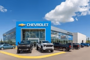 Exterior view of a Chevrolet dealership featuring various pickup trucks parked in front, under a blue sky with fluffy clouds. The dealership building prominently displays the Chevrolet logo.