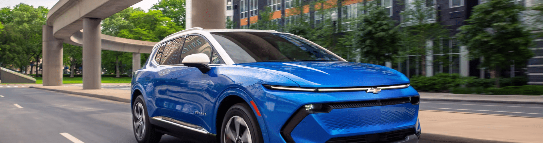 Blue electric SUV driving on a city street with modern architecture and greenery in the background, showcasing its sleek design and innovative features.