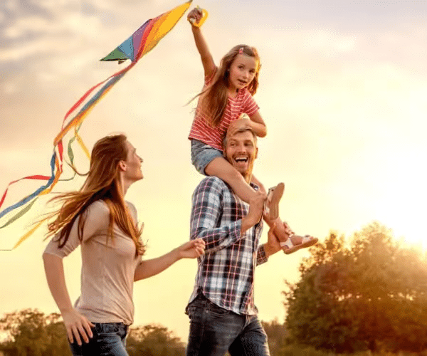 Family enjoying a sunny day outdoors, with a father joyfully carrying his daughter on his shoulders, while the mother walks alongside them. The daughter is holding a colorful kite, and the background features a warm sunset and greenery, capturing the essence of family bonding and outdoor fun.