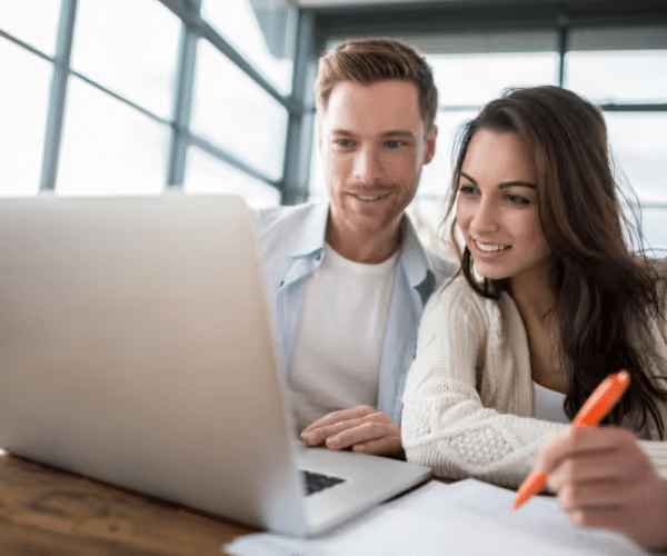 A man and woman collaborate on a laptop, smiling and engaging with the screen while taking notes on paper, in a bright and modern workspace.