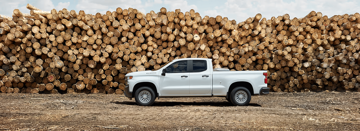 White pickup truck parked in front of a large stack of logs under a blue sky with clouds, showcasing a rural lumber setting suitable for forestry or construction industries.