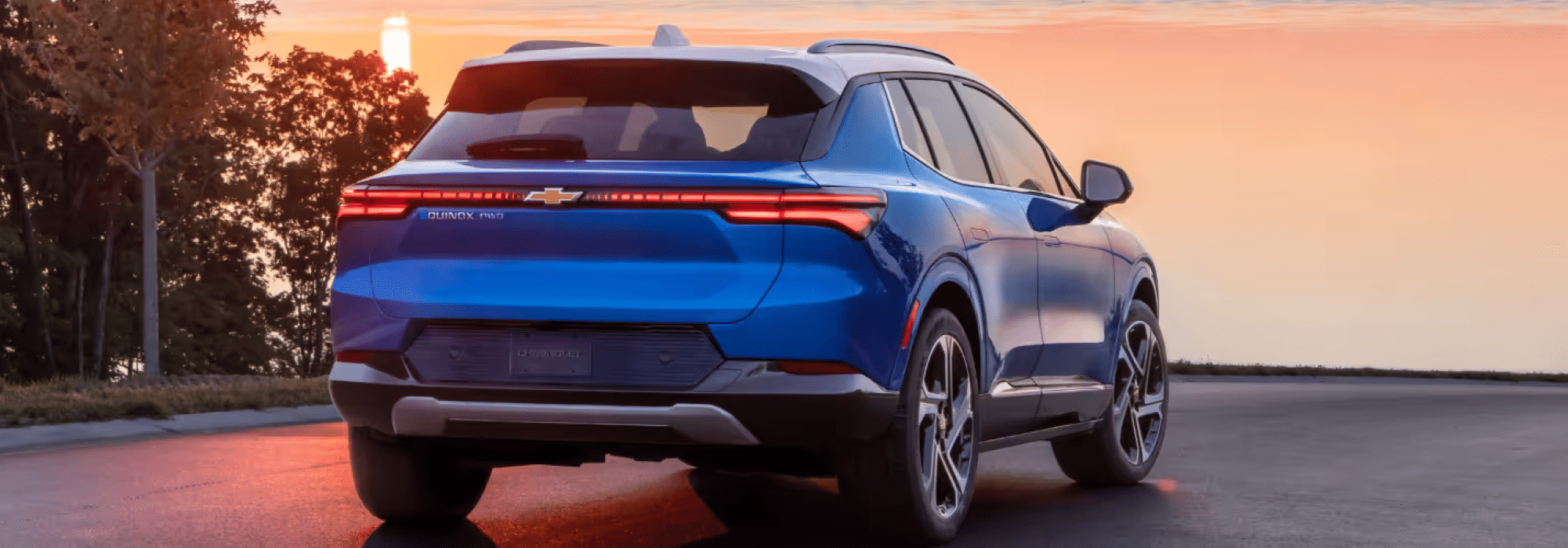 Chevrolet Equinox EV in striking blue color parked on a scenic road at sunset, showcasing its modern rear design, LED taillights, and sleek silhouette against a backdrop of trees and a colorful sky.