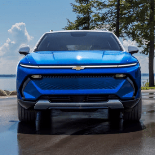 Front view of a blue Chevrolet electric SUV parked near a lake, showcasing its sleek design and modern features under a clear blue sky.