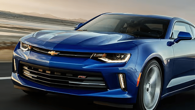 Blue Chevrolet Camaro RS in motion, showcasing its sporty design and sleek lines against a scenic backdrop. The image highlights the car's aggressive front fascia, distinctive headlights, and aerodynamic profile, emphasizing its performance-oriented features.