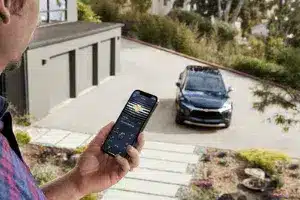 Person using a smartphone to monitor a vehicle parked in a driveway, showcasing smart technology and connectivity in a residential setting.