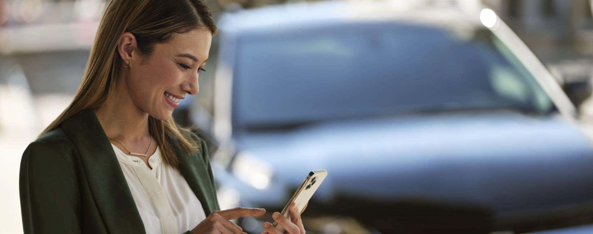 Smiling woman in a green blazer using a smartphone with a car in the background, representing modern technology and urban lifestyle.