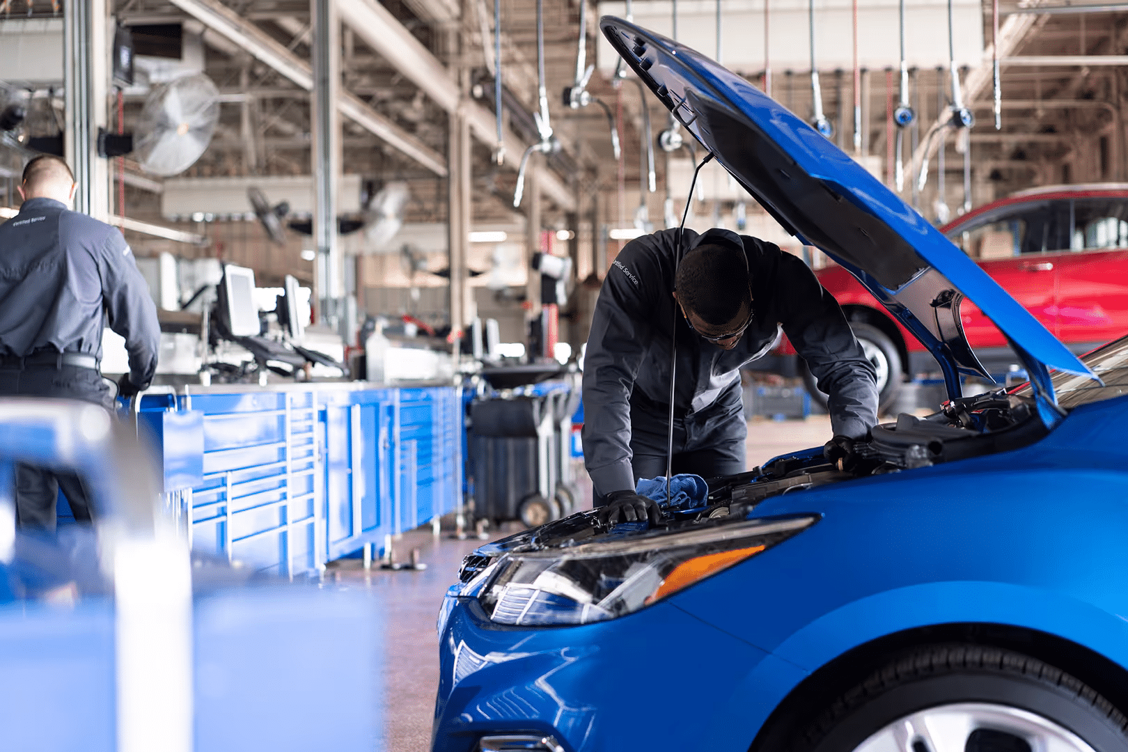 Automobile mechanic performing maintenance on a blue car in an auto repair shop, with tools and equipment visible in the background.