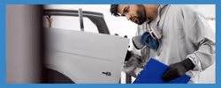 Auto repair technician inspecting a car door panel while holding a clipboard in a workshop setting. The technician is wearing safety gear, including gloves and a face mask, emphasizing safety in automotive repairs.