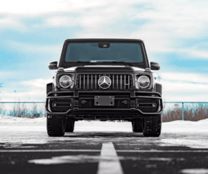Front view of a black Mercedes-Benz G-Class SUV parked in a snowy landscape, showcasing its iconic grille and robust design against a cloudy sky.