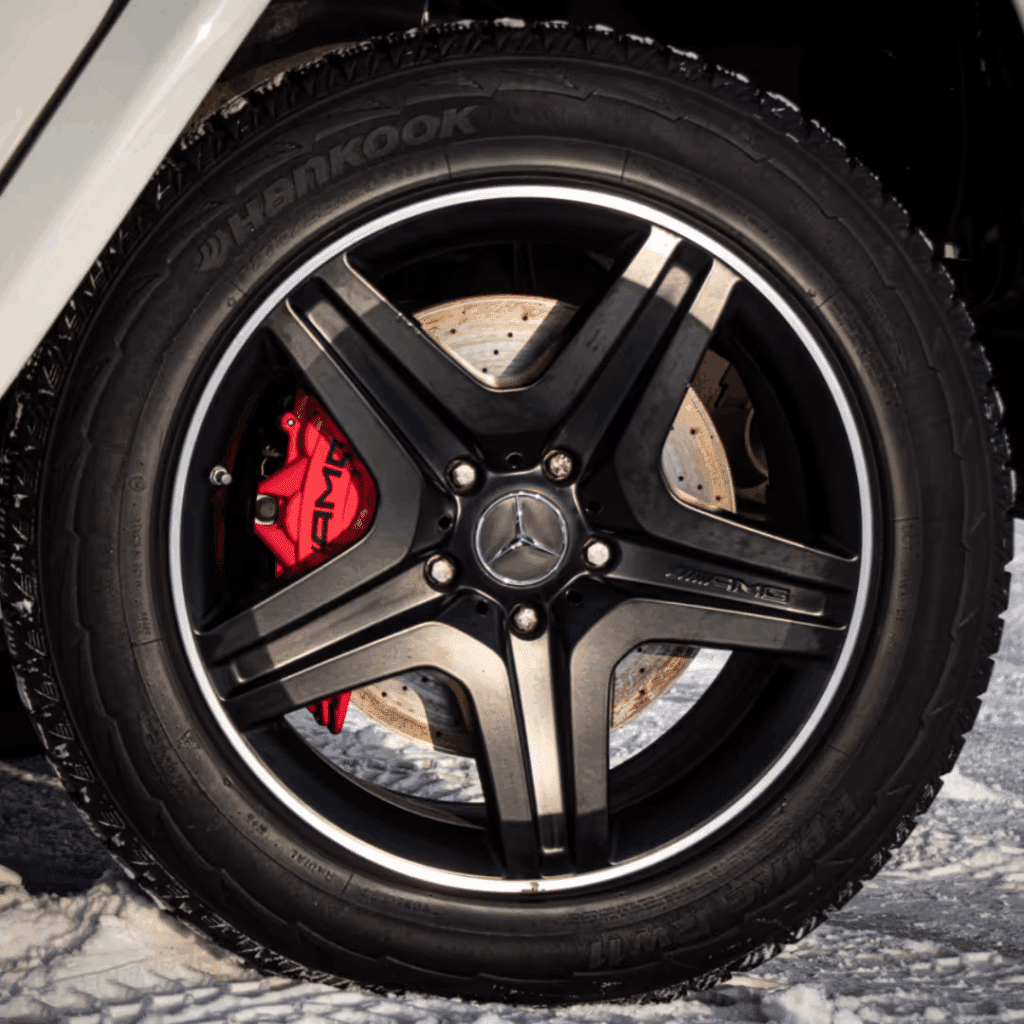 Close-up view of a black alloy wheel with a Mercedes-Benz logo, featuring red brake calipers and Hankook tires, set against a snowy background.
